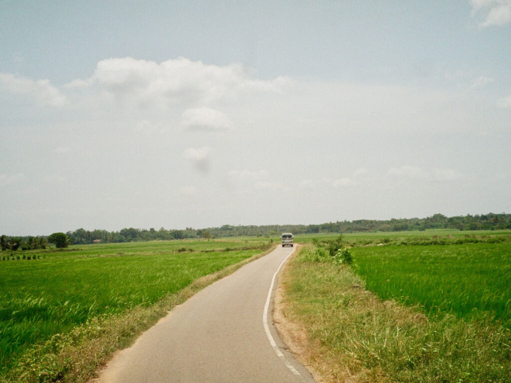 A van on a tarmac road running between bright green paddy fields