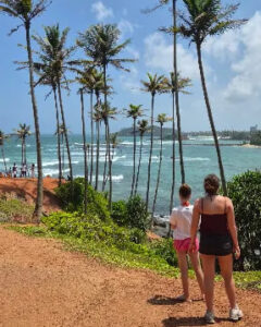 Two travellers on a coastal path under tall coconut palms
