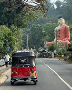 A three-wheeler on a hill road below a large seated Buddha statue